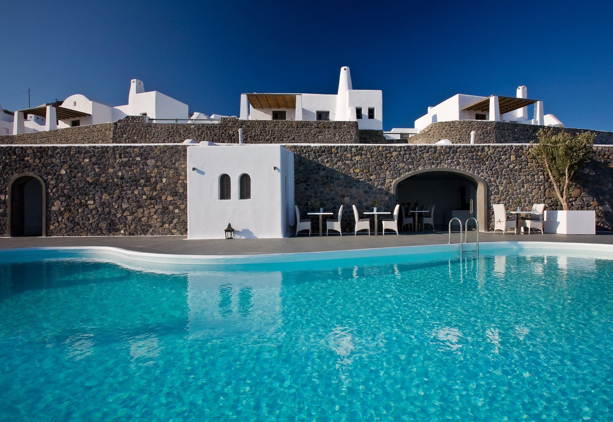 The pool at Carpe Diem Santorini, below whitewashed suites and volcanic-stone terraces under a deep blue sky